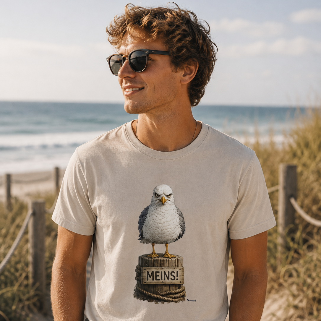Junger Mann trägt das weiße Bio T-Shirt „MEINS!“ mit der Nomavé Möwe am Ostseestrand. Lifestyle-Aufnahme zwischen Dünen mit Blick auf das Meer.