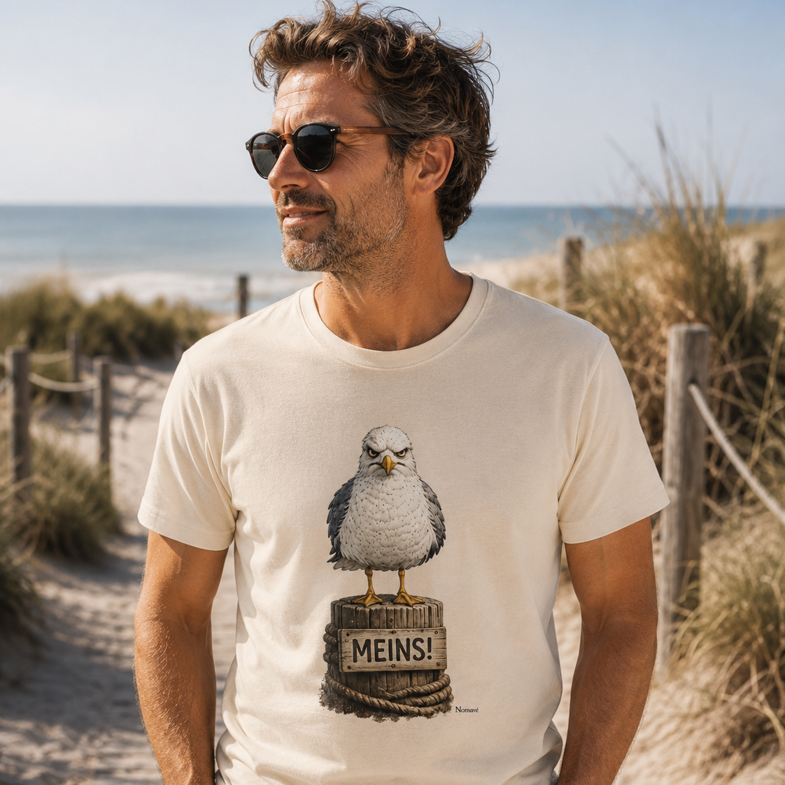 Mann mittleren Alters trägt das weiße Bio T-Shirt „MEINS!“ mit der Nomavé Möwe auf einem Dünenweg am Ostseestrand. Sommerliches Lifestyle-Bild mit Meer im Hintergrund.