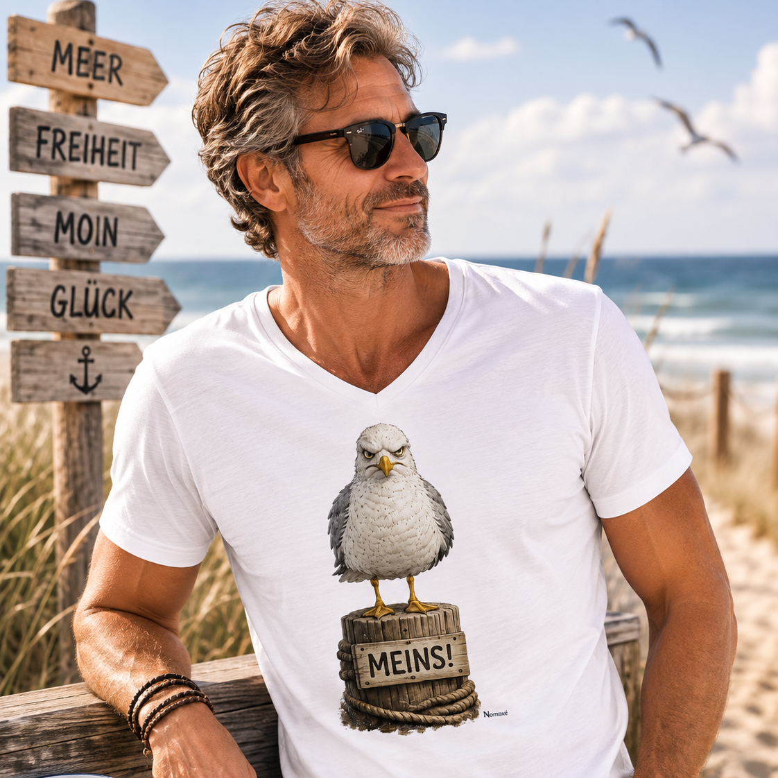 Mann trägt das weiße Bio T-Shirt „MEINS!“ mit der Nomavé Möwe am Ostseestrand. Im Hintergrund Dünen, Meer und ein Wegweiser mit den Worten Meer, Freiheit, Moin und Glück.