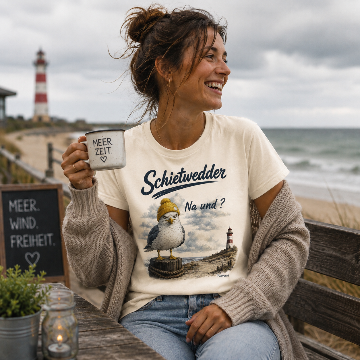 Junge Frau trägt das Nomavé Damen T-Shirt „Schietwedder – Na und?“ mit Möwenmotiv und gelber Mütze an der Ostsee. Mit einer Tasse in der Hand genießt sie den Küstenblick vor Leuchtturm und Meer – gemütlicher norddeutscher Lifestyle.