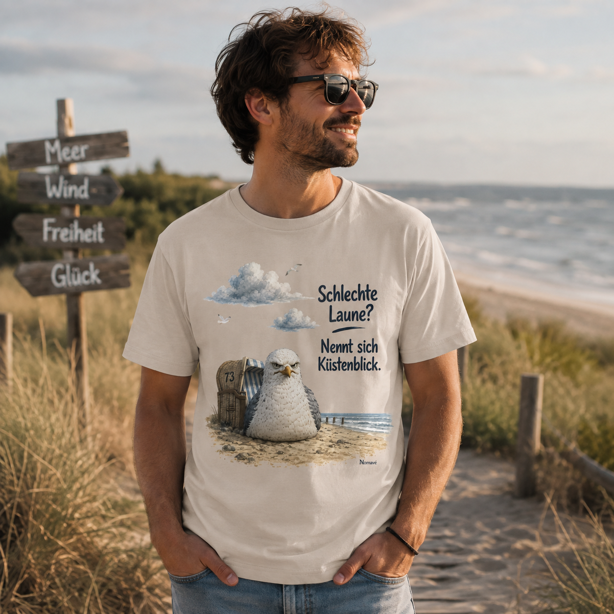 Junger Mann trägt das Nomavé Herren T-Shirt „Schlechte Laune? Nennt sich Küstenblick.“ auf einem Strandweg an der Ostsee. Das maritime Möwenmotiv mit Strandkorb sorgt für einen norddeutschen Küstenlook und entspannten Freizeitstil.