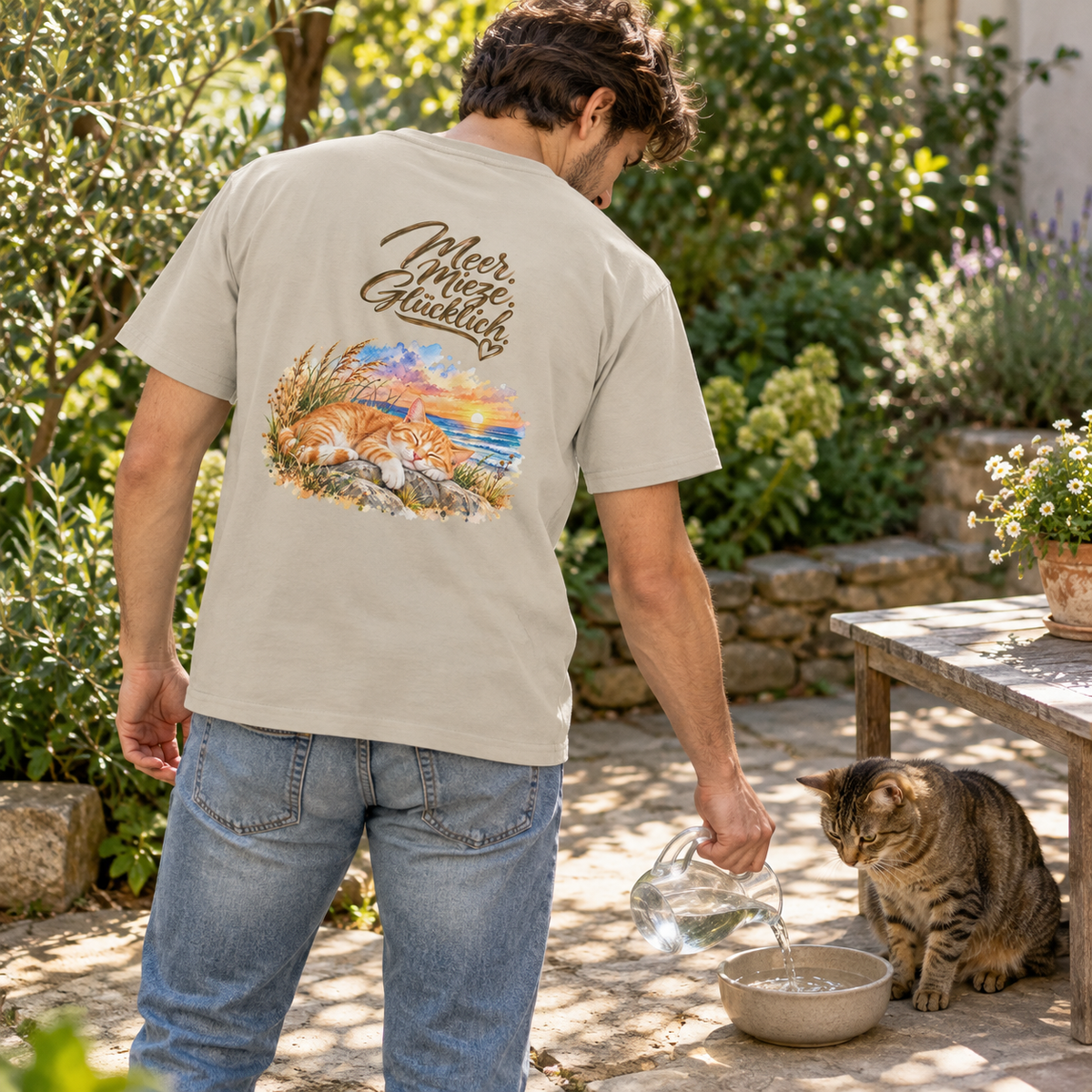 Mann im Heavy-Cotton-T-Shirt in Sand mit „Meer. Mieze. Glücklich.“ als Backprint füllt einer getigerten Katze im Garten Wasser nach.