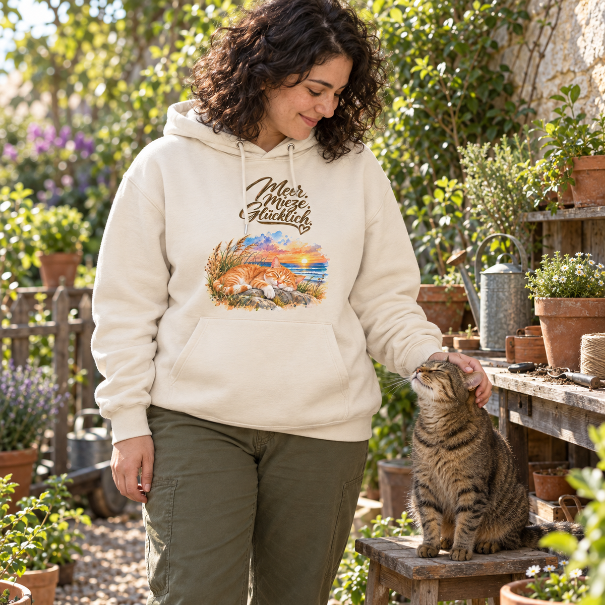 Frau im Bio-Hoodie in Natural Raw mit „Meer. Mieze. Glücklich.“ vorne streichelt eine getigerte Katze im Garten.