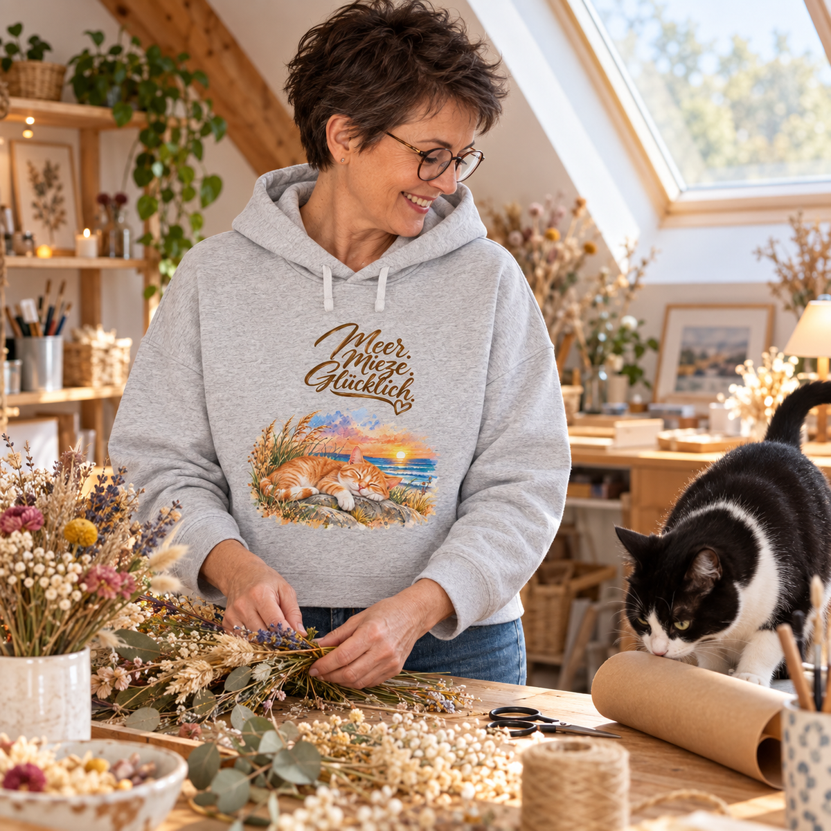 Frau im grauen Damen-Boxcut-Hoodie mit „Meer. Mieze. Glücklich.“ als Frontprint gestaltet Blumen neben einer schwarz-weißen Katze.