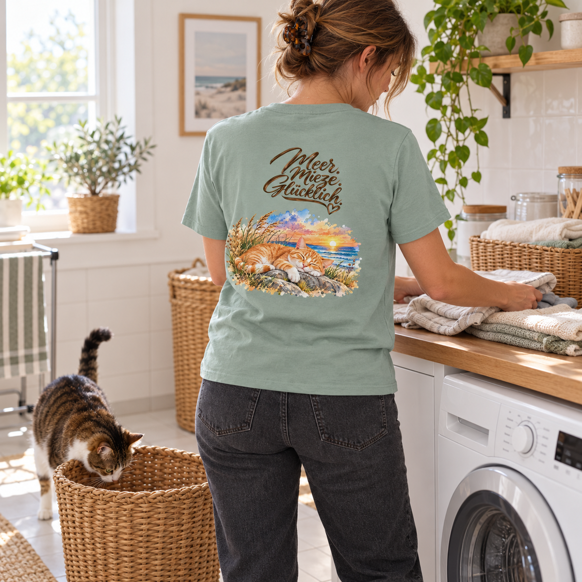 Frau im Bio-T-Shirt in Aloe mit „Meer. Mieze. Glücklich.“ als Backprint faltet Wäsche neben einer Katze am Wäschekorb.