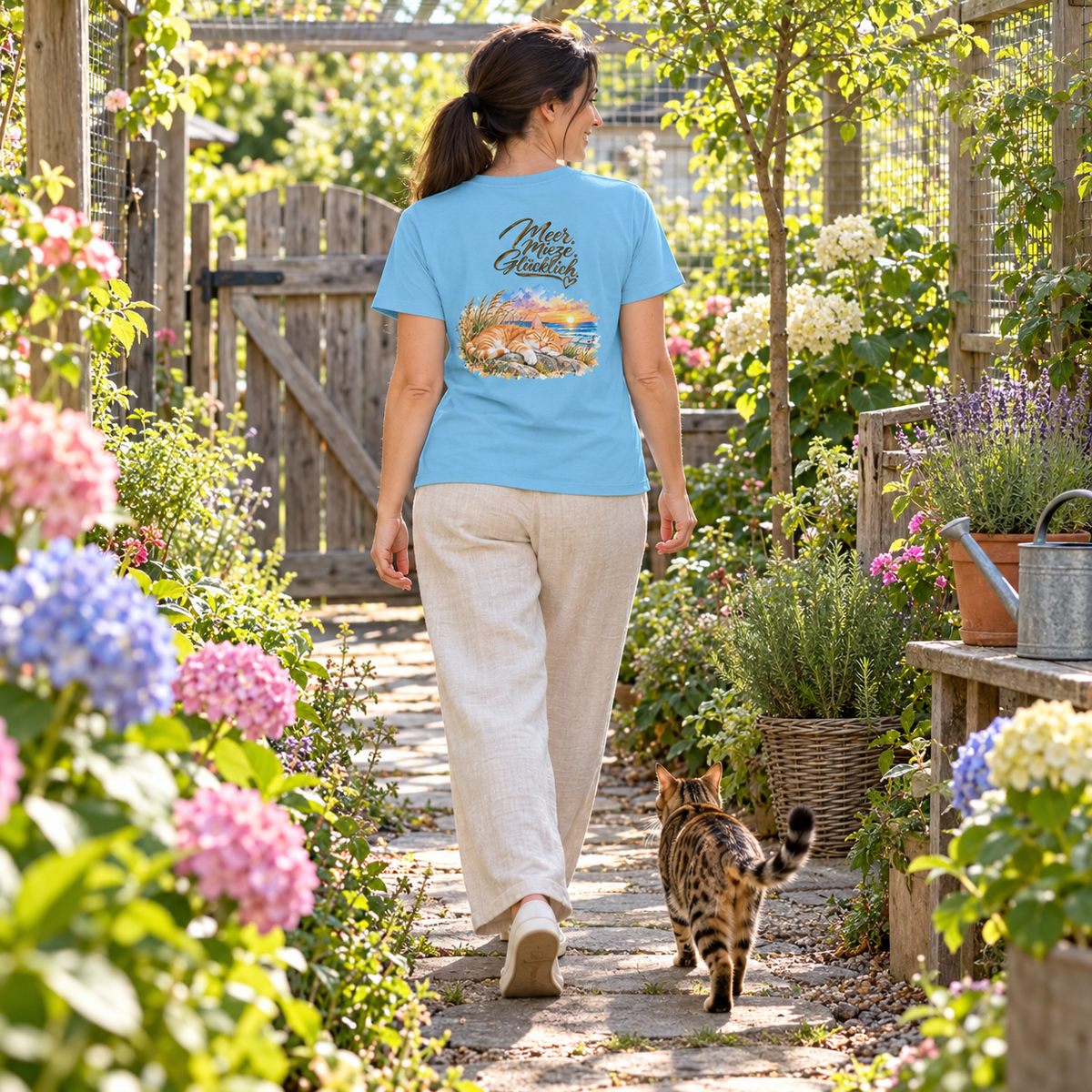Frau im Damen Bio-T-Shirt in Aqua Blue mit „Meer. Mieze. Glücklich.“ auf dem Rücken geht durch einen geschützten Garten; eine getupfte Katze folgt ihr.