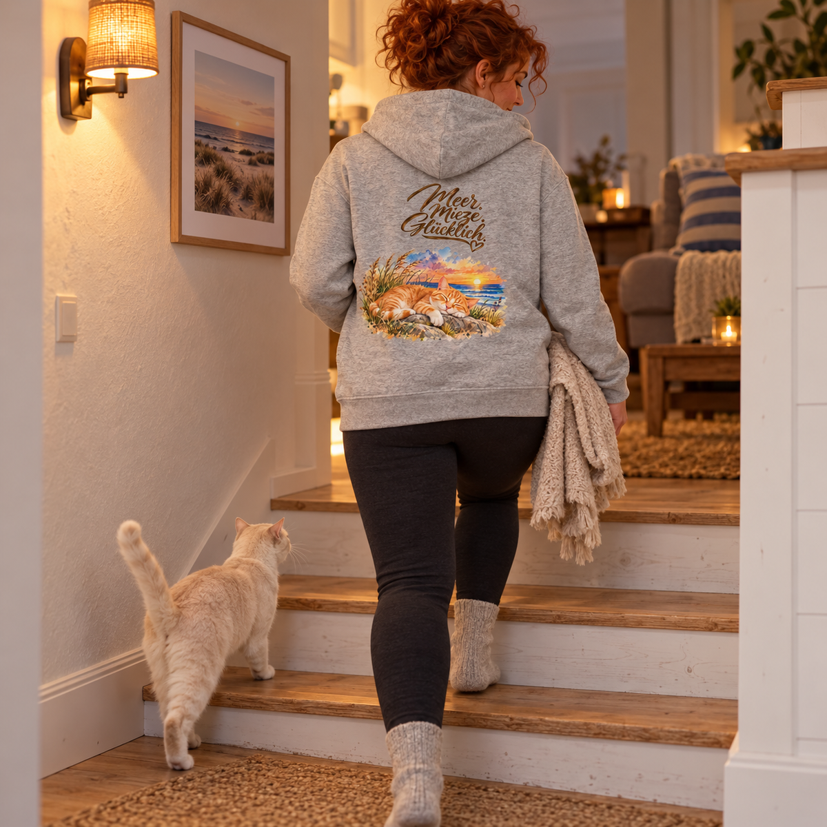 Frau im Bio-Hoodie in Heather Grey mit Katzenmotiv auf dem Rücken trägt eine Decke die Treppe hinauf; eine cremefarbene Katze folgt.
