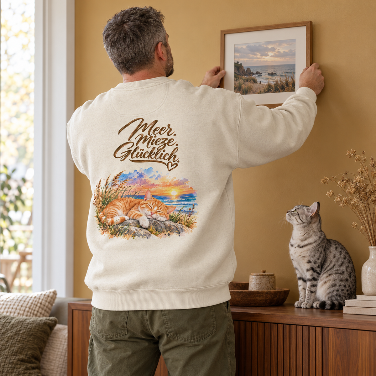 Mann im cremefarbenen Oversize-Sweatshirt mit „Meer. Mieze. Glücklich.“-Katzenmotiv auf dem Rücken hängt ein Küstenbild auf; eine graue Katze sitzt auf der Kommode.