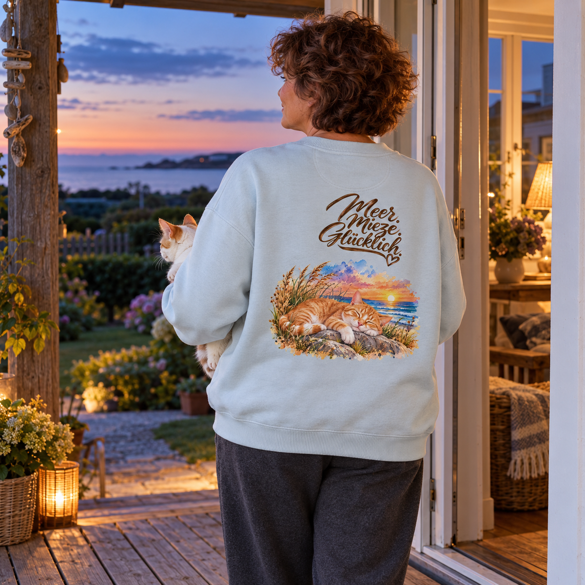 Frau im Oversize-Sweatshirt in Blue Ice mit „Meer. Mieze. Glücklich.“ als Backprint hält eine weiß-orange Katze auf einer Veranda am Abend.