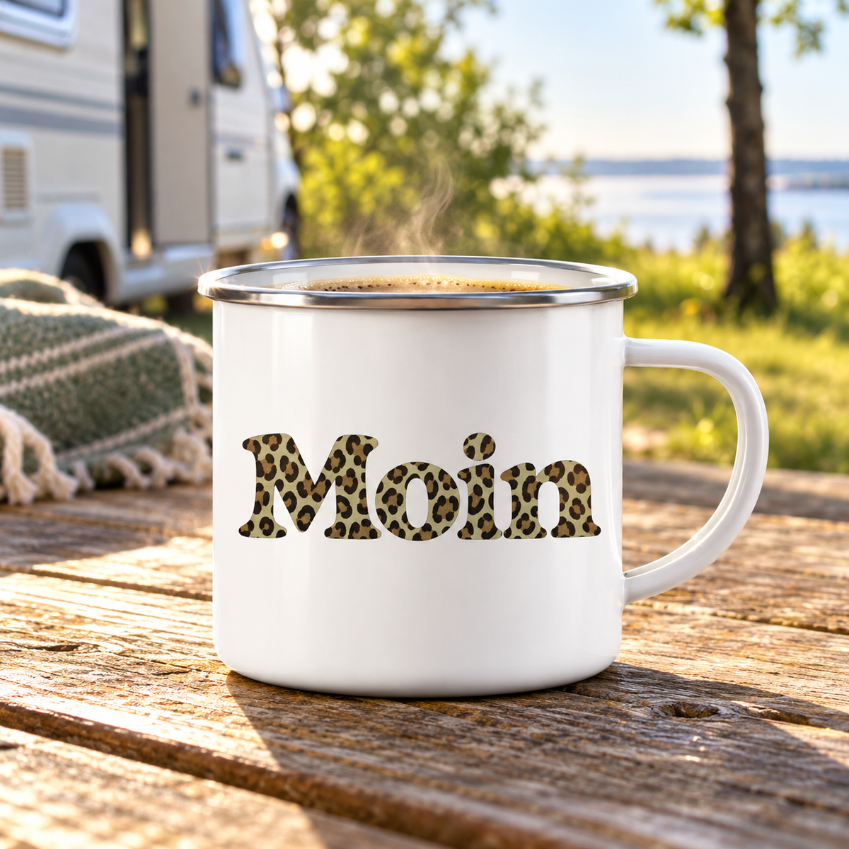 Weiße Emaille-Tasse mit Silberrand und Moin-Leoprint in Sage Green mit Kaffee auf einem Campingtisch.