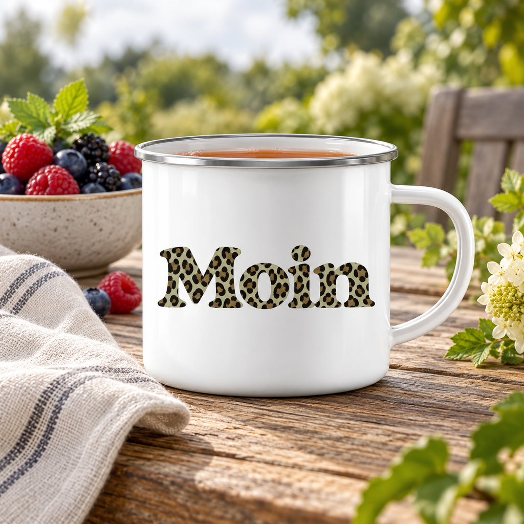 Weiße Emaille-Tasse mit Silberrand und Moin-Leoprint in Sage Green mit Tee auf einem Gartentisch.