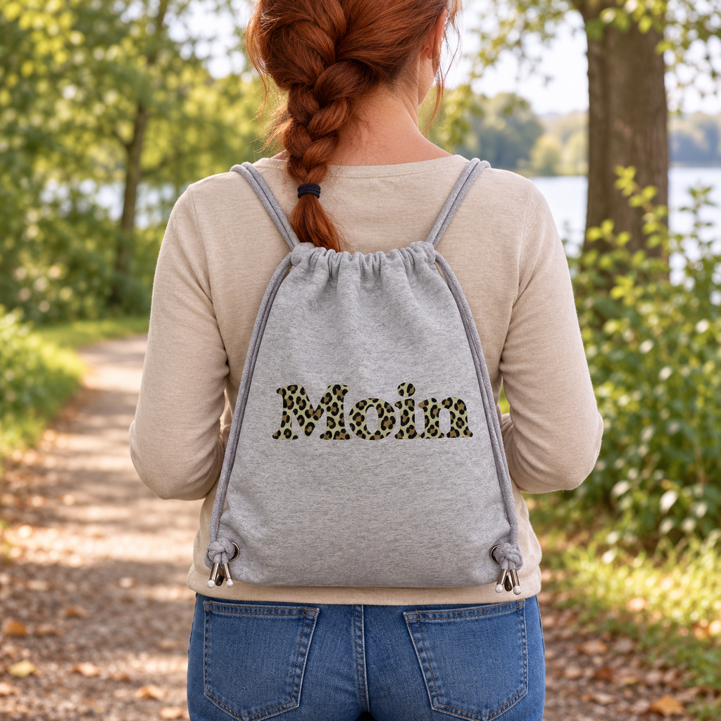 Frau trägt die Gym-Bag in Heather Grey mit Moin-Leoprint in Sage Green auf einem Parkweg, bedruckte Außenseite sichtbar.