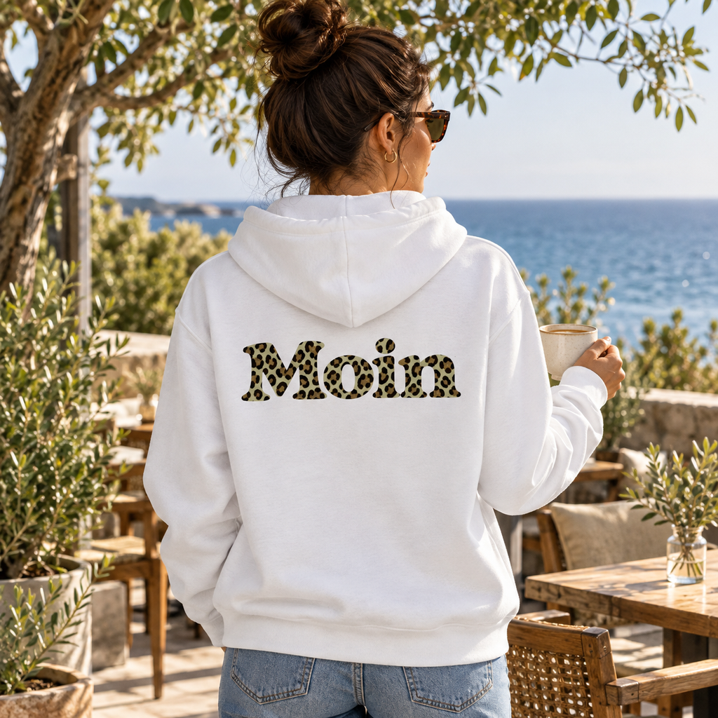 Frau mit weißer Zipper-Kapuzenjacke und Moin-Leo-Sage-Green-Backprint in einem Café.
