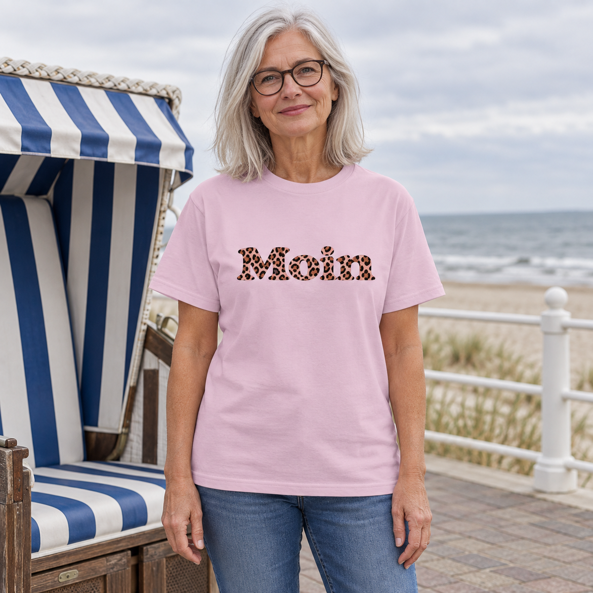 Frau an der Ostseepromenade trägt ein Heavy-Cotton-T-Shirt in Light Pink mit Moin-Leo-Blush-Pink-Motiv.
