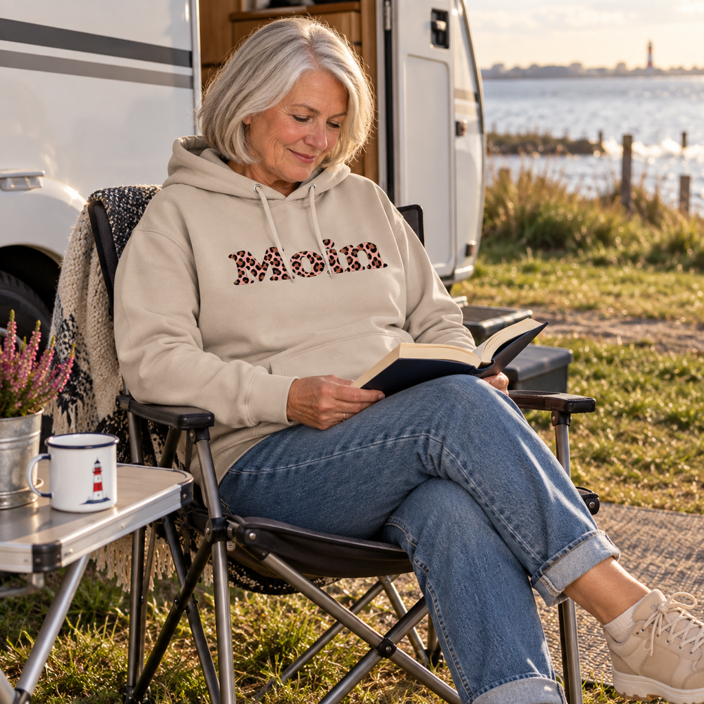 Frau liest am Camper im Bio-Unisex-Hoodie in Desert Dust mit Moin-Leo-Blush-Pink-Frontprint.