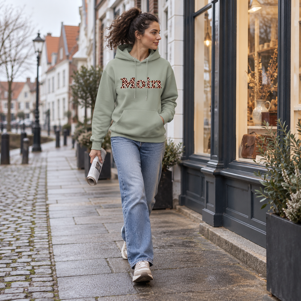 Gehende Frau trägt den Bio-Unisex-Hoodie in Aloe mit Moin-Leo-Blush-Pink-Motiv.