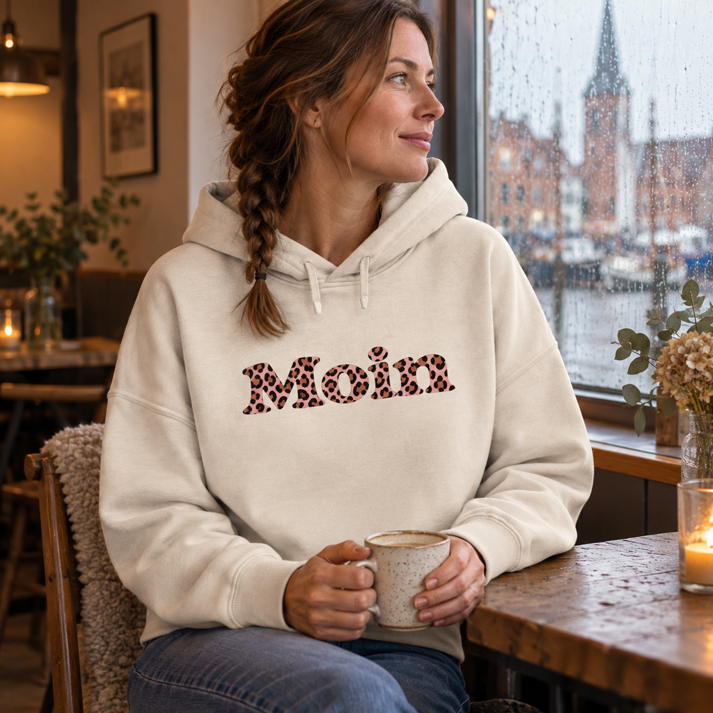 Frau sitzt im Café im Damen-Bio-Boxcut-Hoodie in Natural Raw mit Moin-Leo-Blush-Pink-Frontprint.