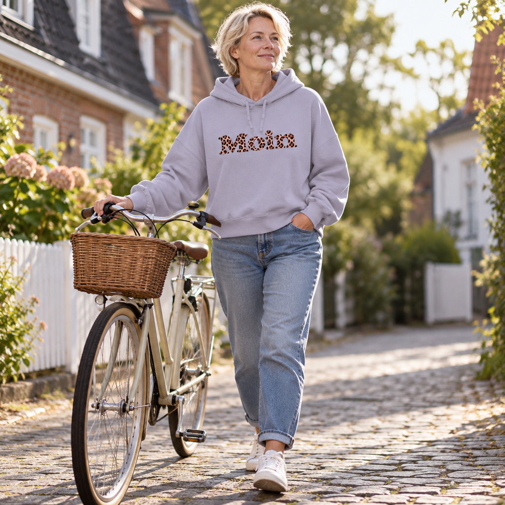 Frau schiebt ein Fahrrad im Damen-Bio-Boxcut-Hoodie in Lavender mit Moin-Leo-Blush-Pink-Motiv.