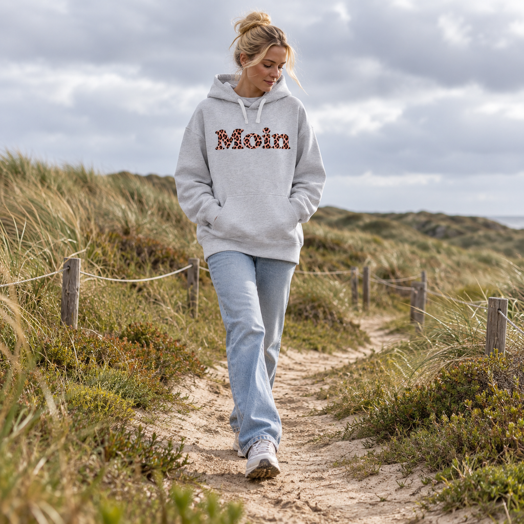 Frau spaziert auf einem Dünenweg im Bio-Oversize-Hoodie in Cool Heather Grey mit Moin-Leo-Blush-Pink-Frontprint.