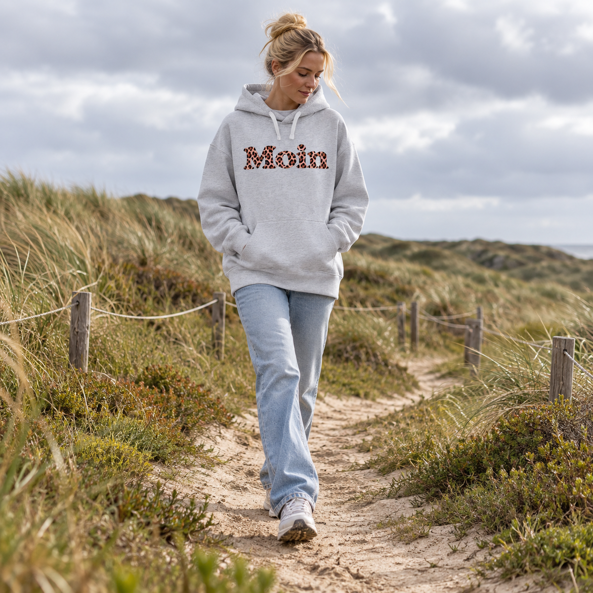 Frau spaziert auf einem Dünenweg im Bio-Oversize-Hoodie in Cool Heather Grey mit Moin-Leo-Blush-Pink-Frontprint.