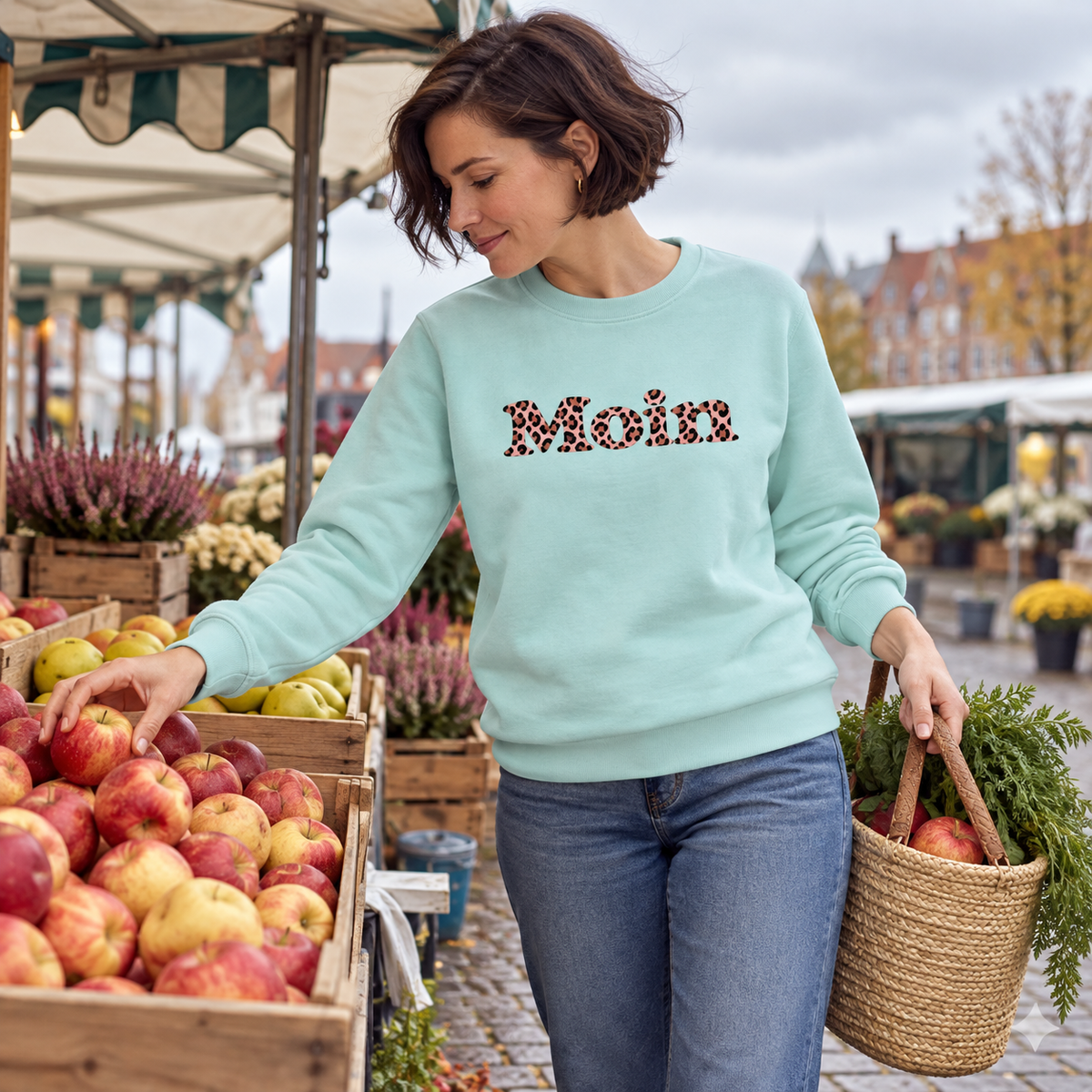 Frau wählt Äpfel auf dem Wochenmarkt im Bio-Unisex-Sweatshirt Caribbean Blue mit rosafarbenem Moin-Leo-Frontprint.