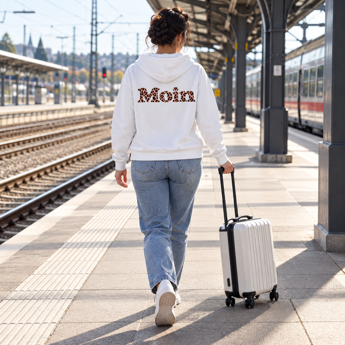 Frau geht mit Koffer auf einem Bahnsteig, weiße Bio-Zipper-Kapuzenjacke mit sichtbarem Moin-Leo-Blush-Pink-Backprint.