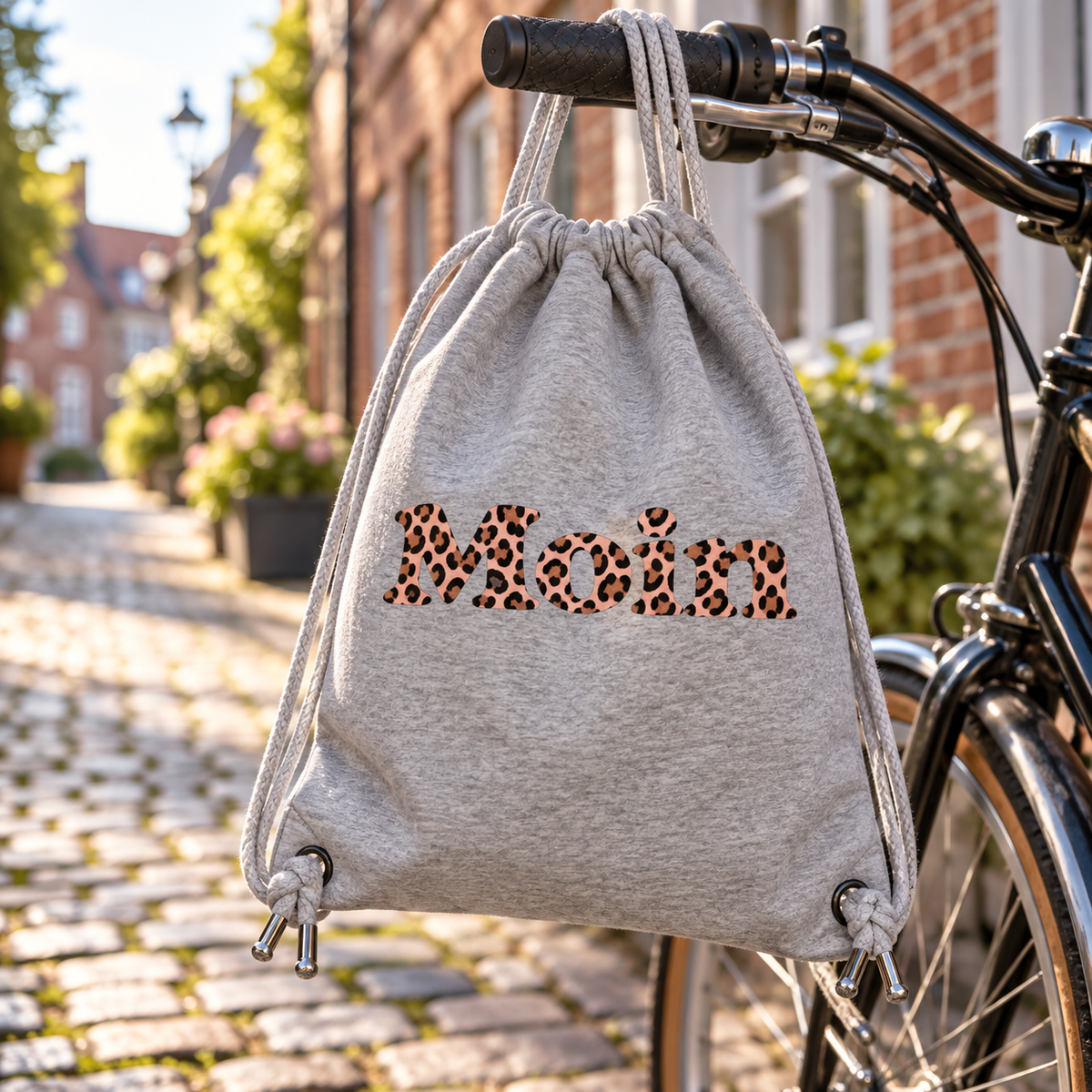 Gym-Bag Heather Grey mit Moin Leo Blush Pink hängt am Fahrradlenker in einer gepflasterten Straße.