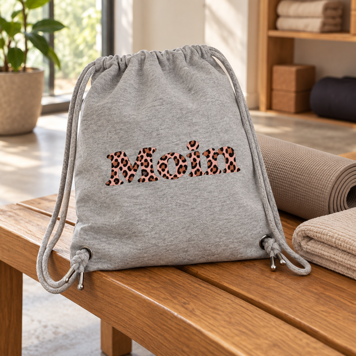 Gym-Bag Heather Grey mit rosafarbenem Moin-Leo-Motiv steht neben Yogamatte und Handtuch auf einer Holzbank.