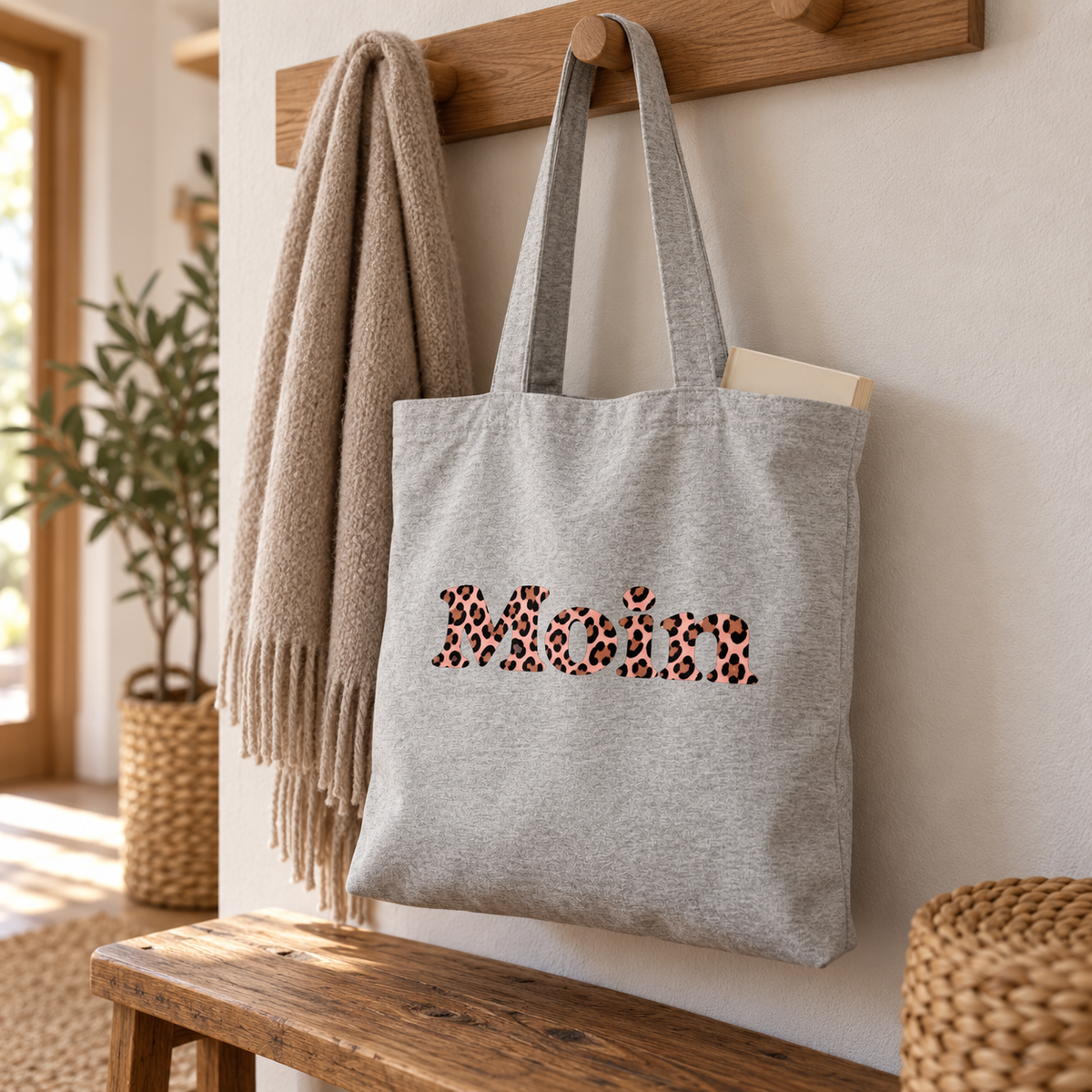 Tote-Bag Heather Grey mit Moin-Leo-Blush-Pink-Motiv hängt an einer Garderobe neben einem Schal.