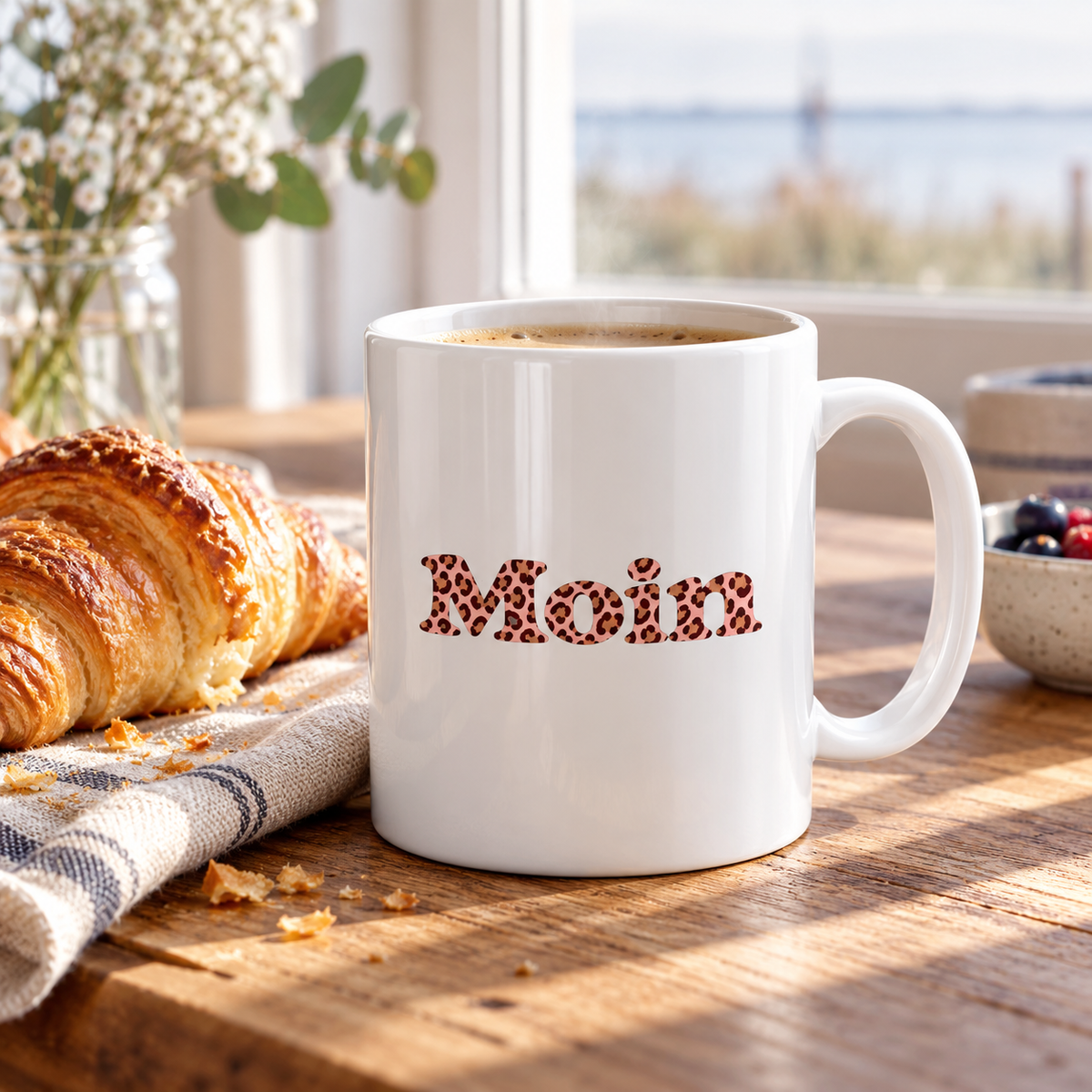 Weiße Glossy-Tasse mit Moin Leo Blush Pink und Kaffee neben einem Croissant auf dem Frühstückstisch.