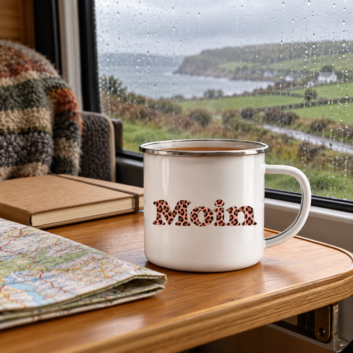 Emaille-Tasse mit Silberrand und Moin-Leo-Blush-Pink-Motiv auf einem Campertisch neben Karte und Notizbuch.