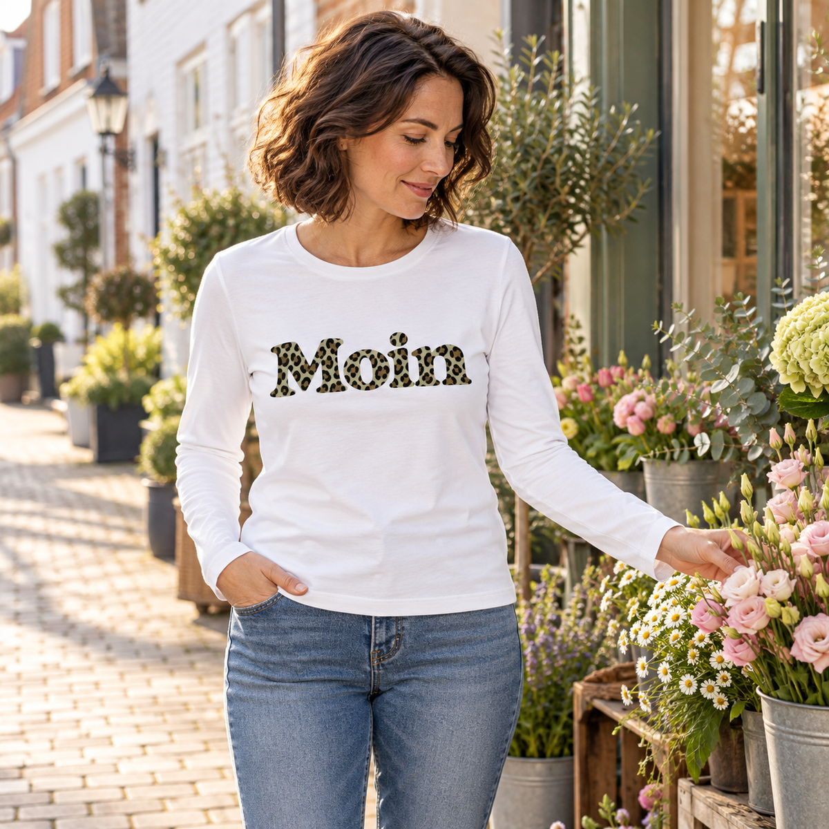 Frau im weißen Damen Bio Longsleeve mit Moin-Leoprint in Sage Green vor einem Blumenladen, Vorderansicht.