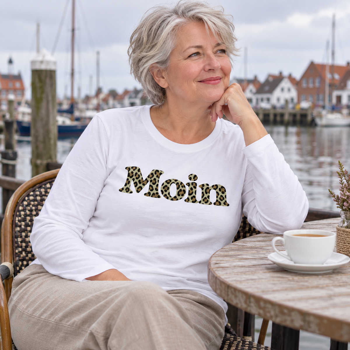 Frau im weißen Damen Bio Longsleeve mit Moin-Leoprint in Sage Green an einem Hafencafé, Vorderansicht.