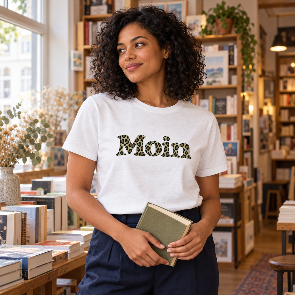 Frau im weißen Damen Bio T-Shirt mit Moin-Leoprint in Sage Green in einer Buchhandlung, Vorderansicht.