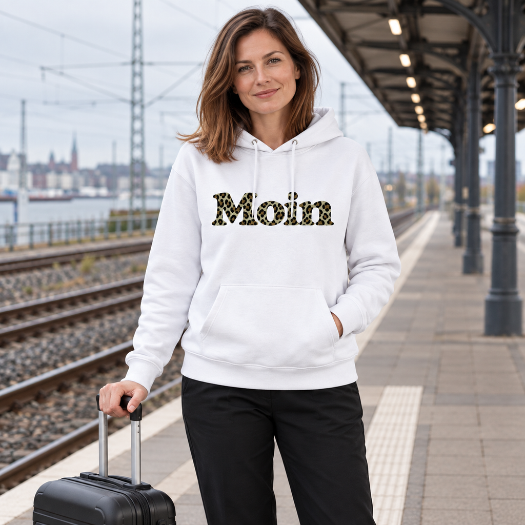 Frau im weißen Bio Unisex Hoodie mit Moin-Leoprint in Sage Green auf einem Bahnsteig, Vorderansicht.