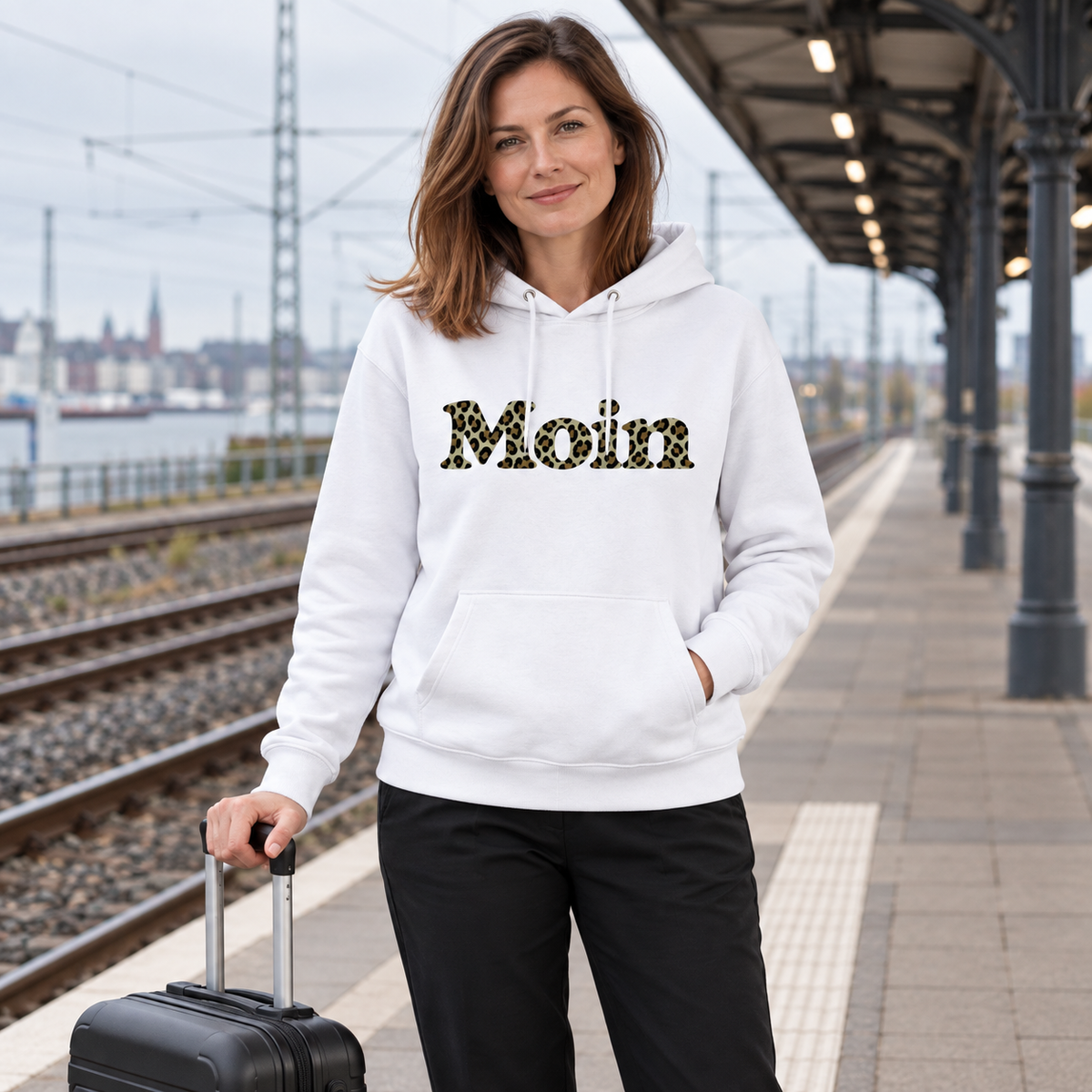 Frau im weißen Bio Unisex Hoodie mit Moin-Leoprint in Sage Green auf einem Bahnsteig, Vorderansicht.