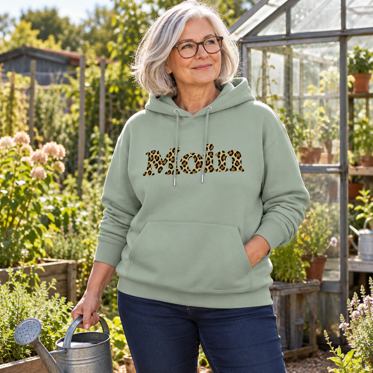 Frau im Bio Unisex Hoodie in Aloe mit Moin-Leoprint in Sage Green im Garten, Vorderansicht.