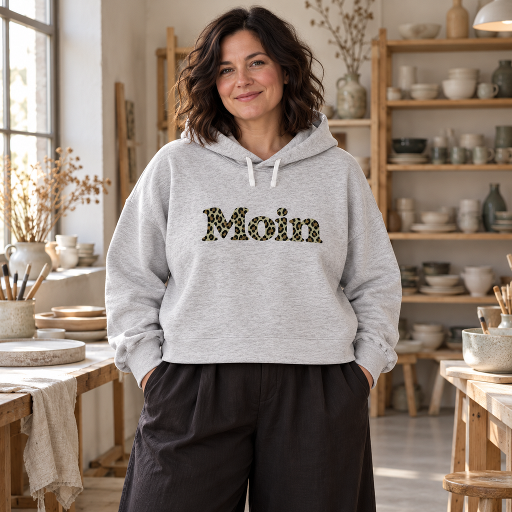 Frau im Damen Bio Boxcut Hoodie in Cool Heather Grey mit Moin-Leoprint in Sage Green in einem Keramikatelier, Vorderansicht.