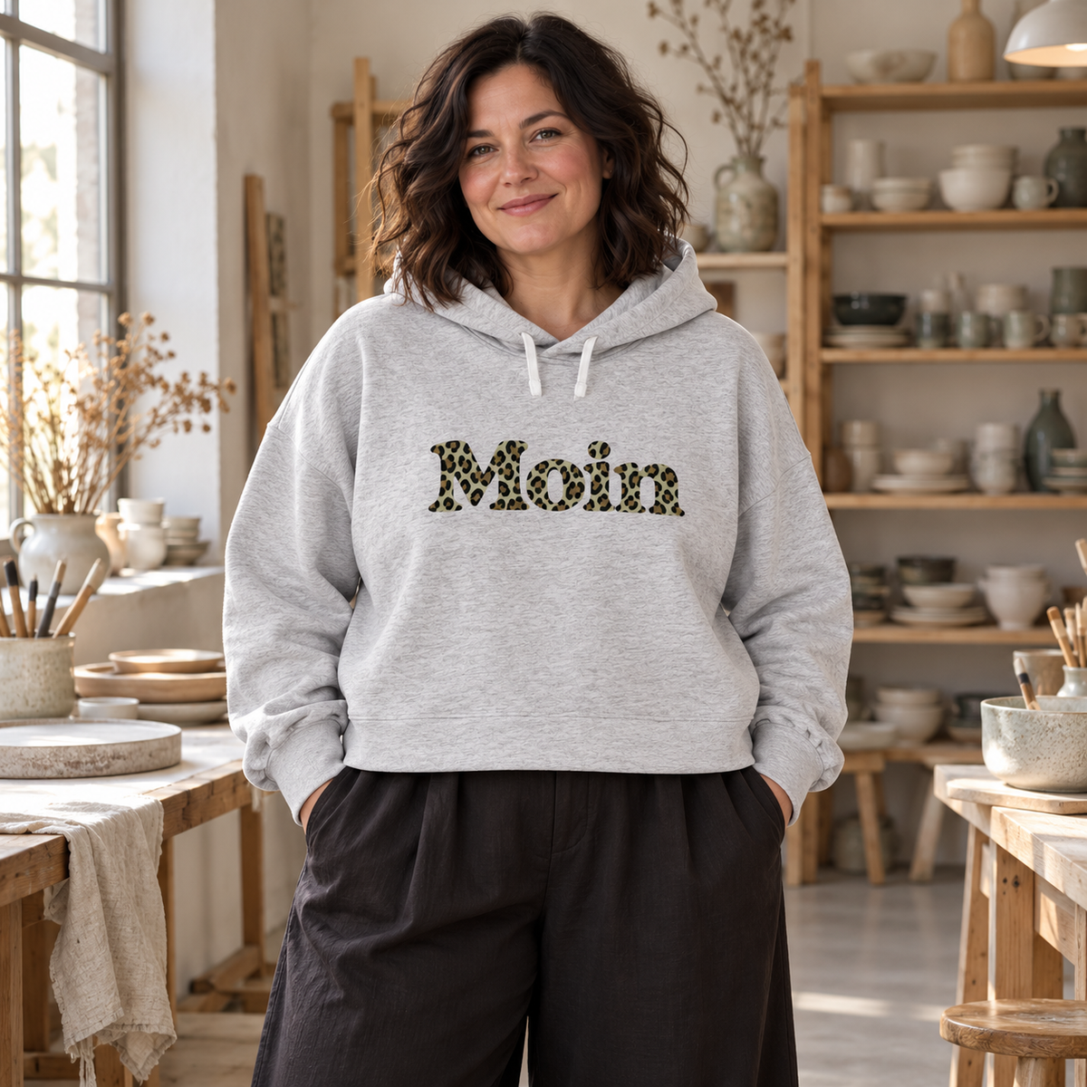 Frau im Damen Bio Boxcut Hoodie in Cool Heather Grey mit Moin-Leoprint in Sage Green in einem Keramikatelier, Vorderansicht.