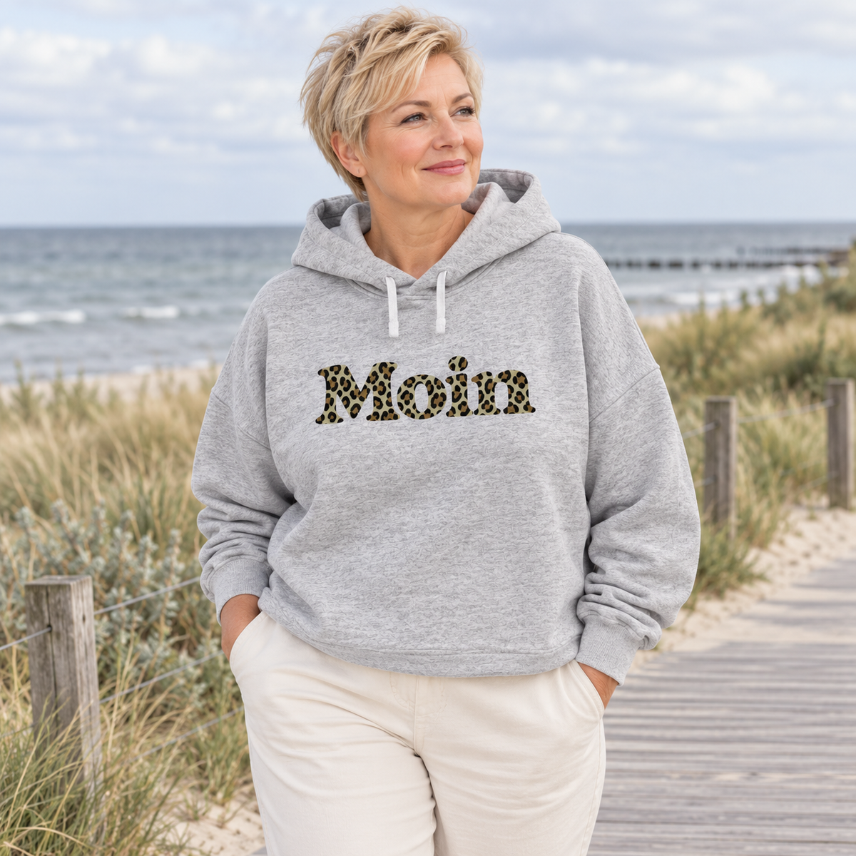 Frau im Damen Bio Boxcut Hoodie in Cool Heather Grey mit Moin-Leoprint in Sage Green auf einer Küstenpromenade, Vorderansicht.