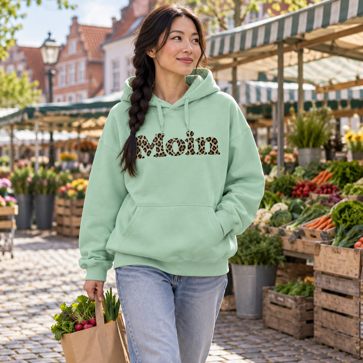 Frau im Bio Oversize Hoodie in Misty Jade mit Moin-Leoprint in Sage Green auf einem Wochenmarkt, Vorderansicht.