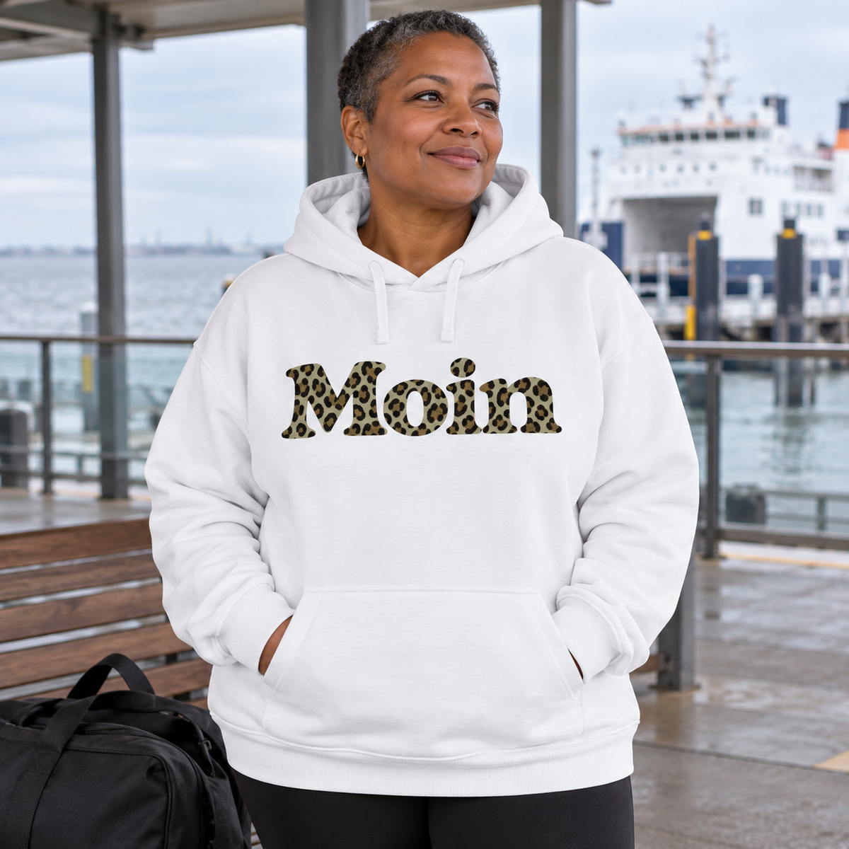 Frau im weißen Bio Oversize Hoodie mit Moin-Leoprint in Sage Green an einem Fährterminal, Vorderansicht.