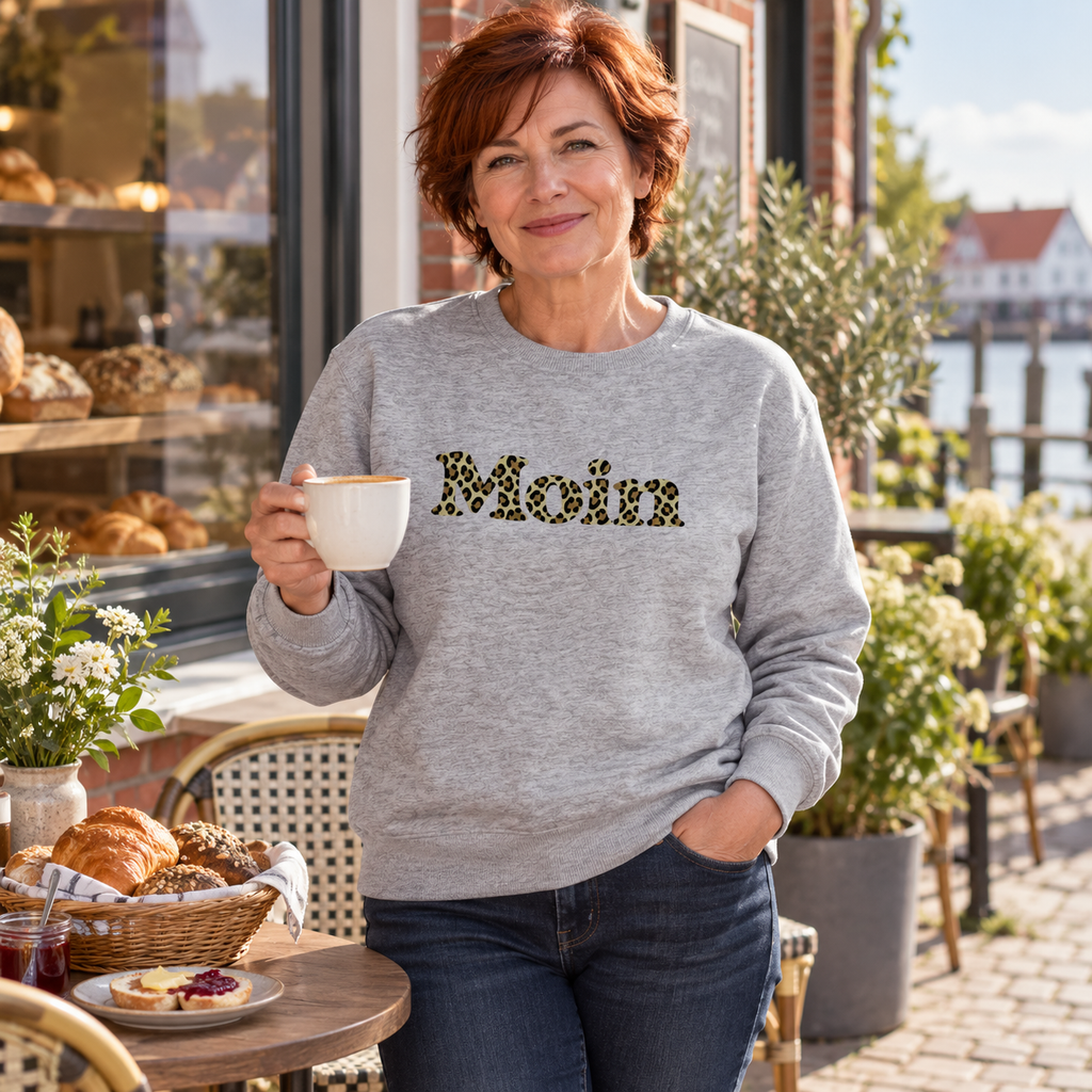 Frau im Bio Unisex Sweatshirt in Heather Grey mit Moin-Leoprint in Sage Green auf einer Bäckereiterrasse, Vorderansicht.