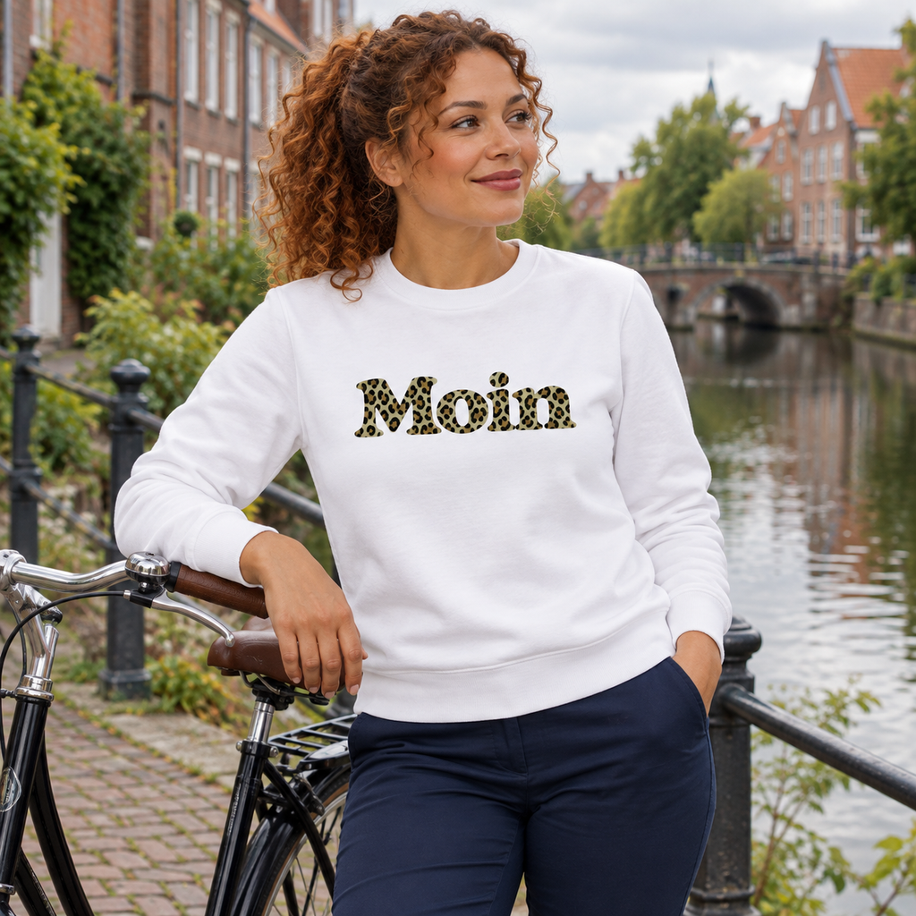 Frau im weißen Bio Unisex Sweatshirt mit Moin-Leoprint in Sage Green neben einem Fahrrad am Kanal, Vorderansicht.