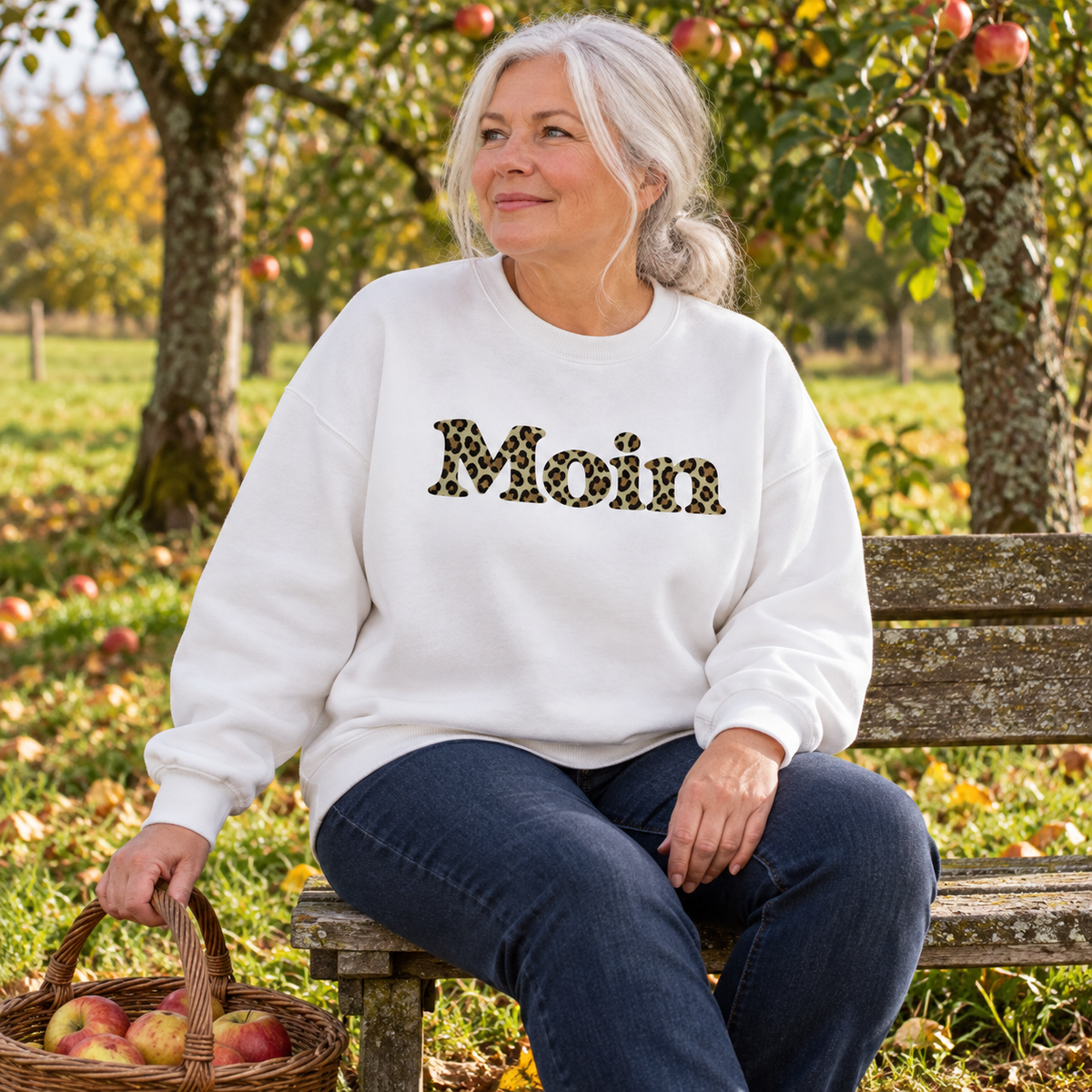 Frau im weißen Bio Oversize Sweatshirt mit Moin-Leoprint in Sage Green auf einer Bank im Obstgarten, Vorderansicht.