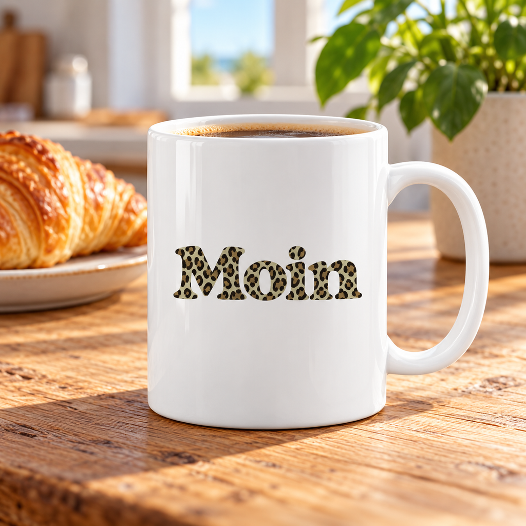 Weiße glänzende Tasse mit Moin-Leoprint in Sage Green und Kaffee auf einem Frühstückstisch, Henkel rechts.