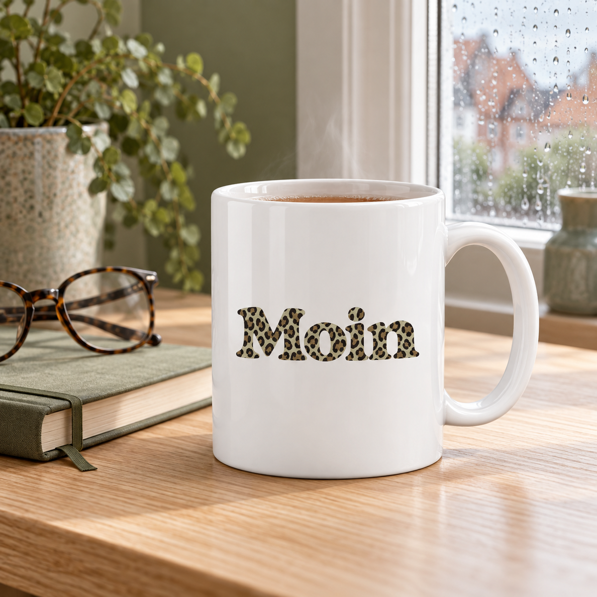 Weiße glänzende Tasse mit Moin-Leoprint in Sage Green und Tee neben einem Notizbuch am Fenster, Henkel rechts.