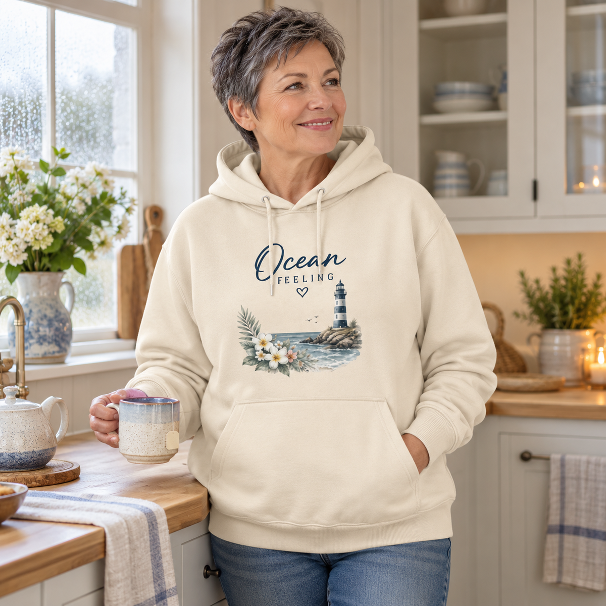 Frau im Ocean Feeling Unisex Bio-Hoodie in Natural Raw mit floralem Frontprint beim Tee in einer Ferienwohnung