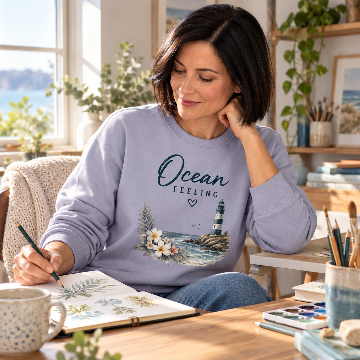 Frau im Ocean Feeling Unisex Bio-Sweatshirt in Serene Blue mit Blumen und Leuchtturm vorne beim Zeichnen zu Hause