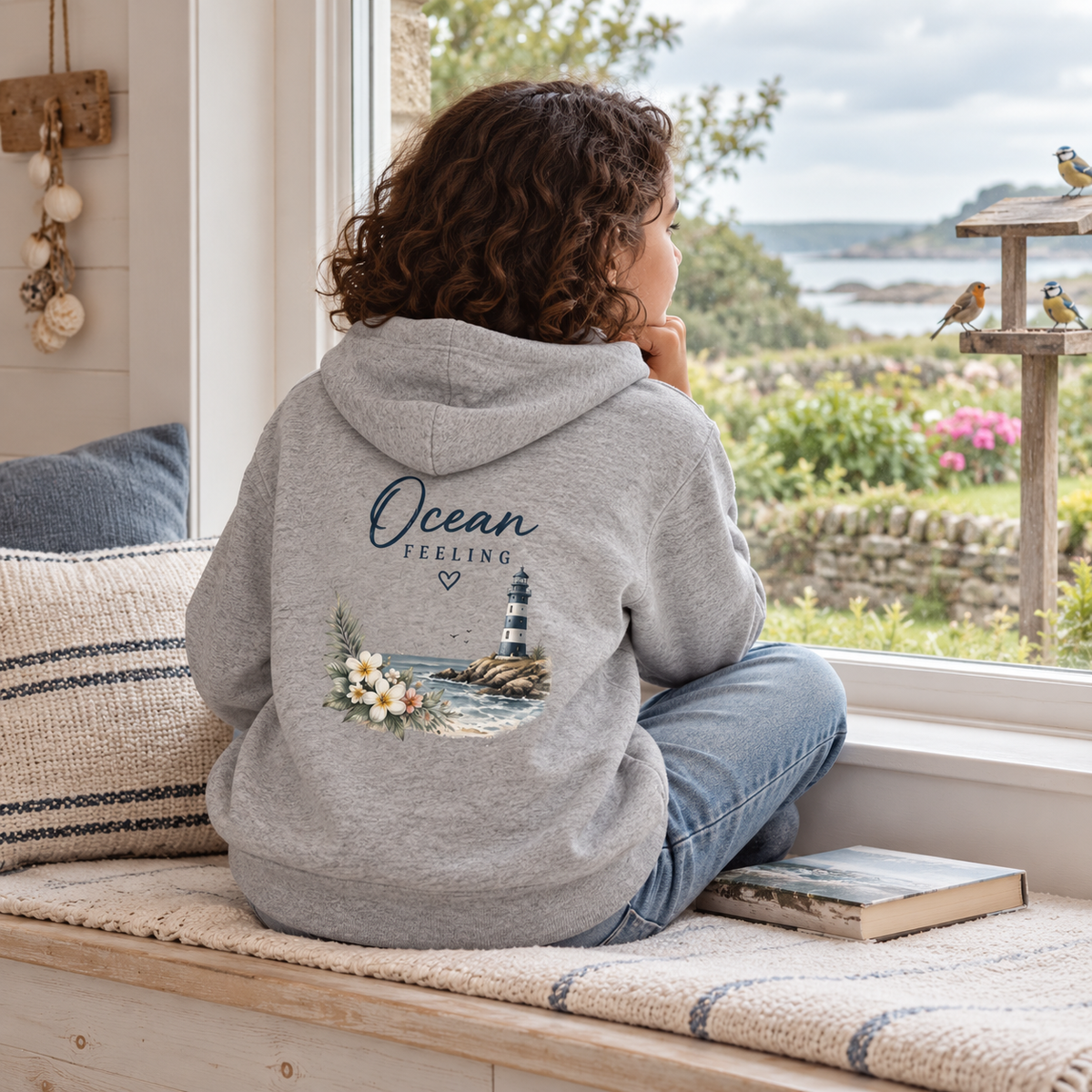 Mädchen im Ocean Feeling Kinder Bio-Hoodie in Heather Grey mit Blumen und Leuchtturm auf dem Rücken am Fenster